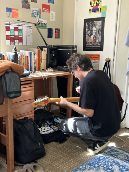 Gallis crouched at a desk, holding a guitar, back turned to the camera