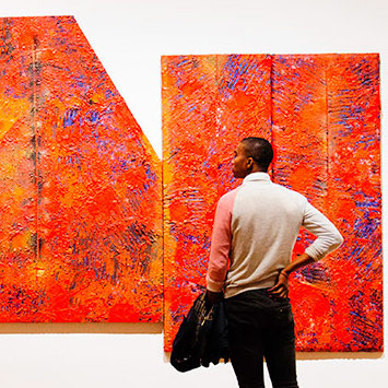 A student stands in front of a bright red abstract painting, his back to the viewer
