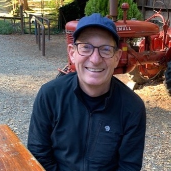 A photo of Aaron Straight wearing a ball cap, sitting at a picnic table with a red tractor in the background