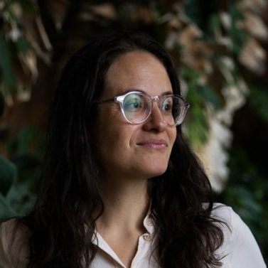 Person with long brown hair and glasses glancing diagonally with natural elements in the background.