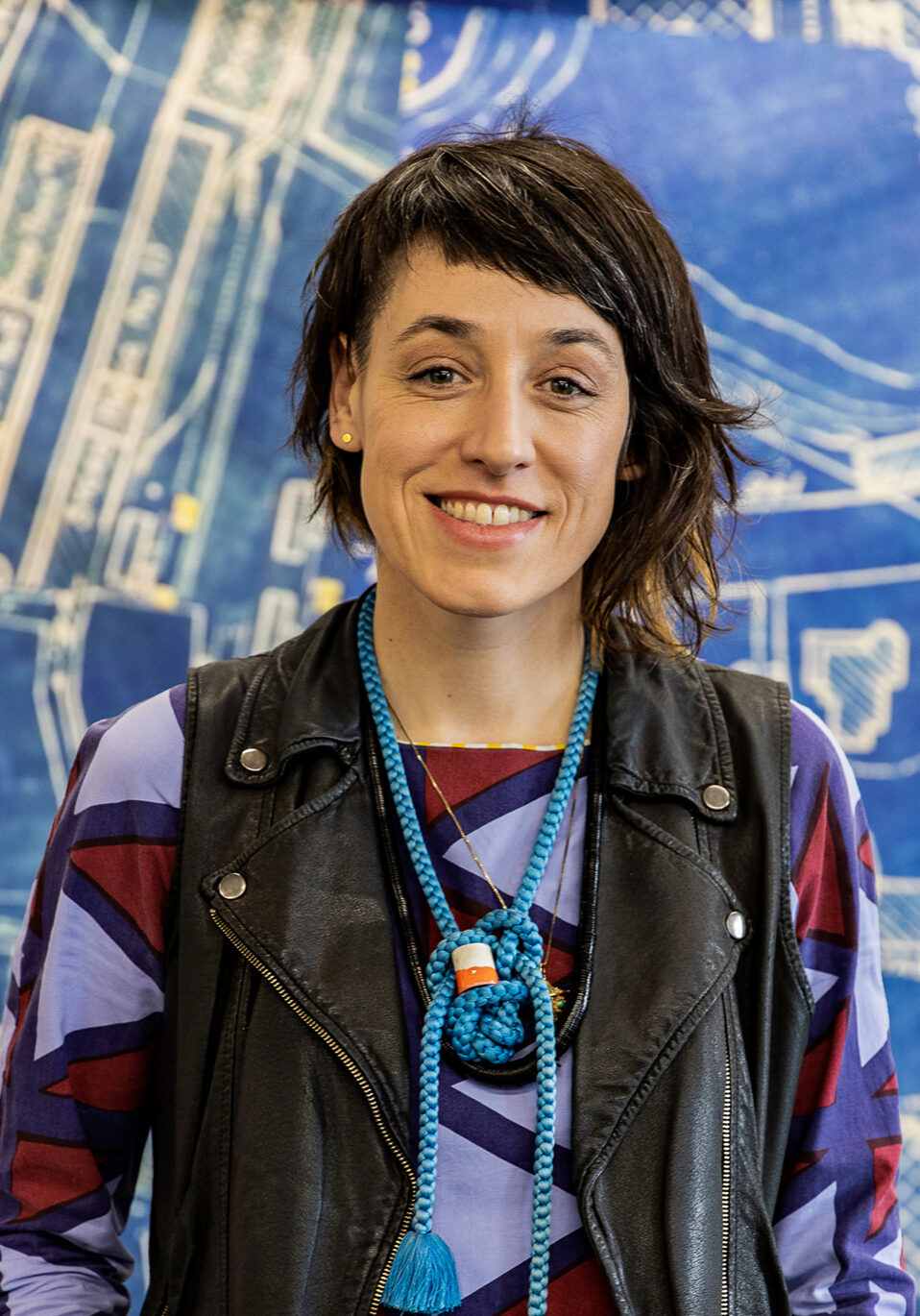 A photo of Amanda Curreri, a person with shoulder-length dark hair, wearing a blue and black patterned shirt and standing in front of a blue and white painted background