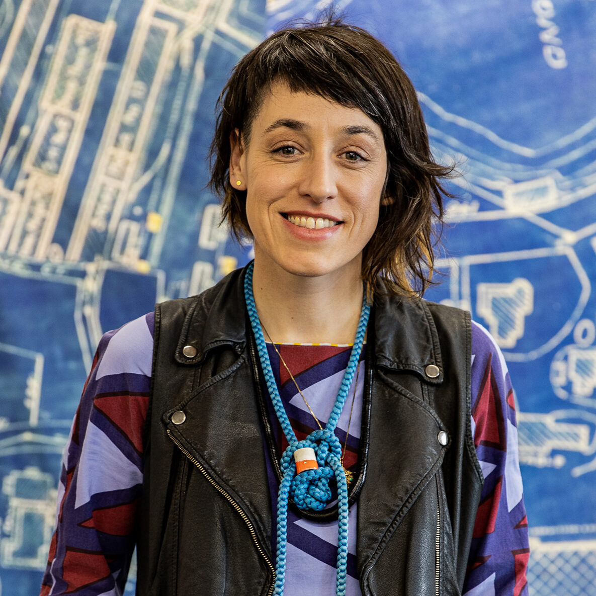 A photo of Amanda Curreri, a person with shoulder-length dark hair, wearing a blue and black patterned shirt and standing in front of a blue and white painted background