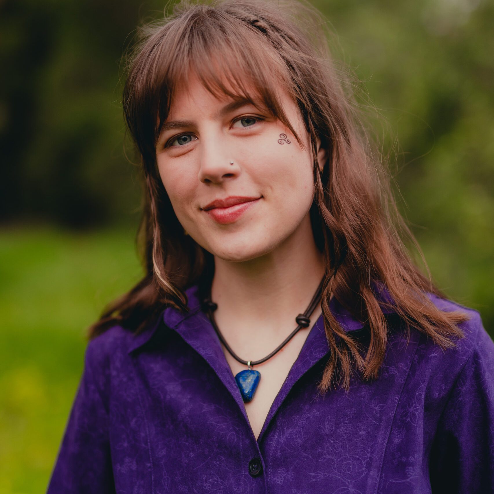 Portrait of Camilla Wickman: a young white woman with a stone necklace, in front of green foliage