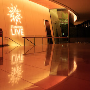 Curved lobby of Bing Concert Hall illuminated by ceiling lights. The Stanford Live sunburst logo is projected on a wall.