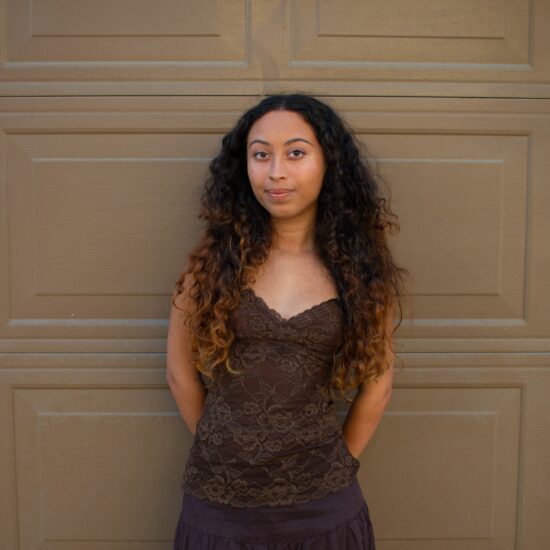 A photo of Tristyn dressed in brown, leaning against a brown wall