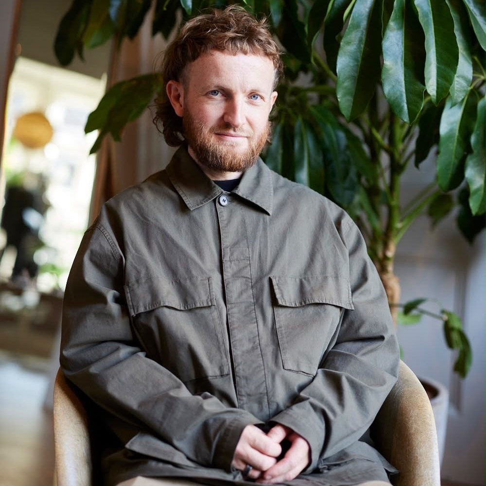 Person in grey button up shirt sitting in a chair in front of an indoor plant and mirror