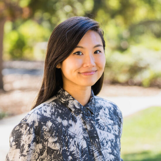 Edi Dai, person with long dark brown hair and black and white button up shirt in front of lush green background