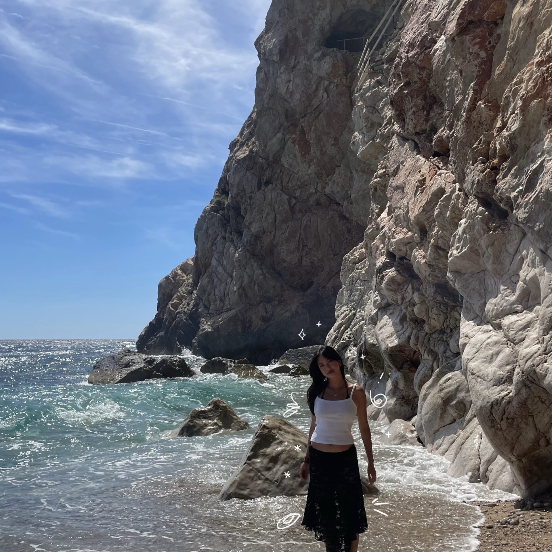 A photo of Stella standing with her feet in the ocean near a rocky cliff