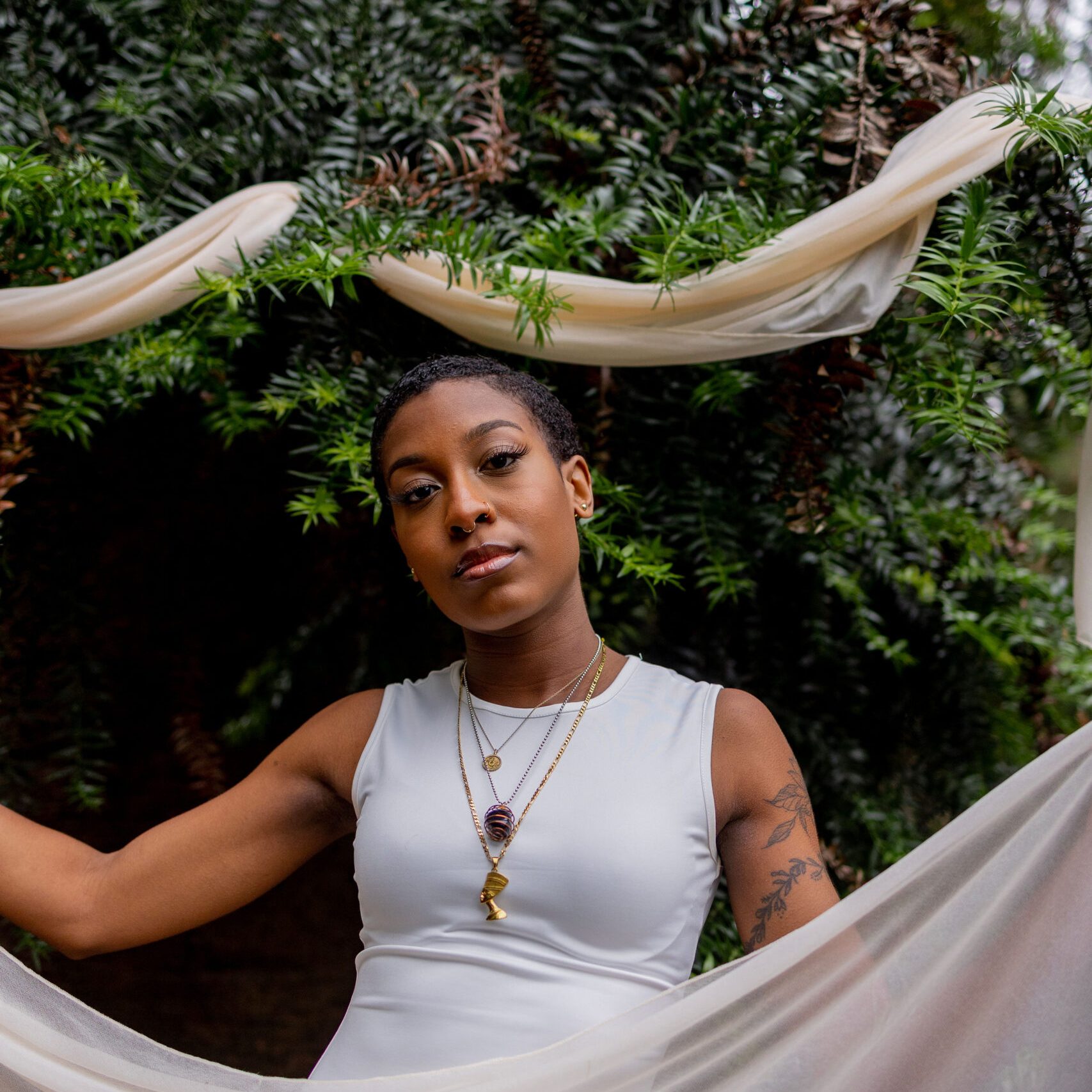 Portrait of Tyah-Amoy "Yoma" Roberts: a young black woman in a white dress with beige flowy material in front of green foliage