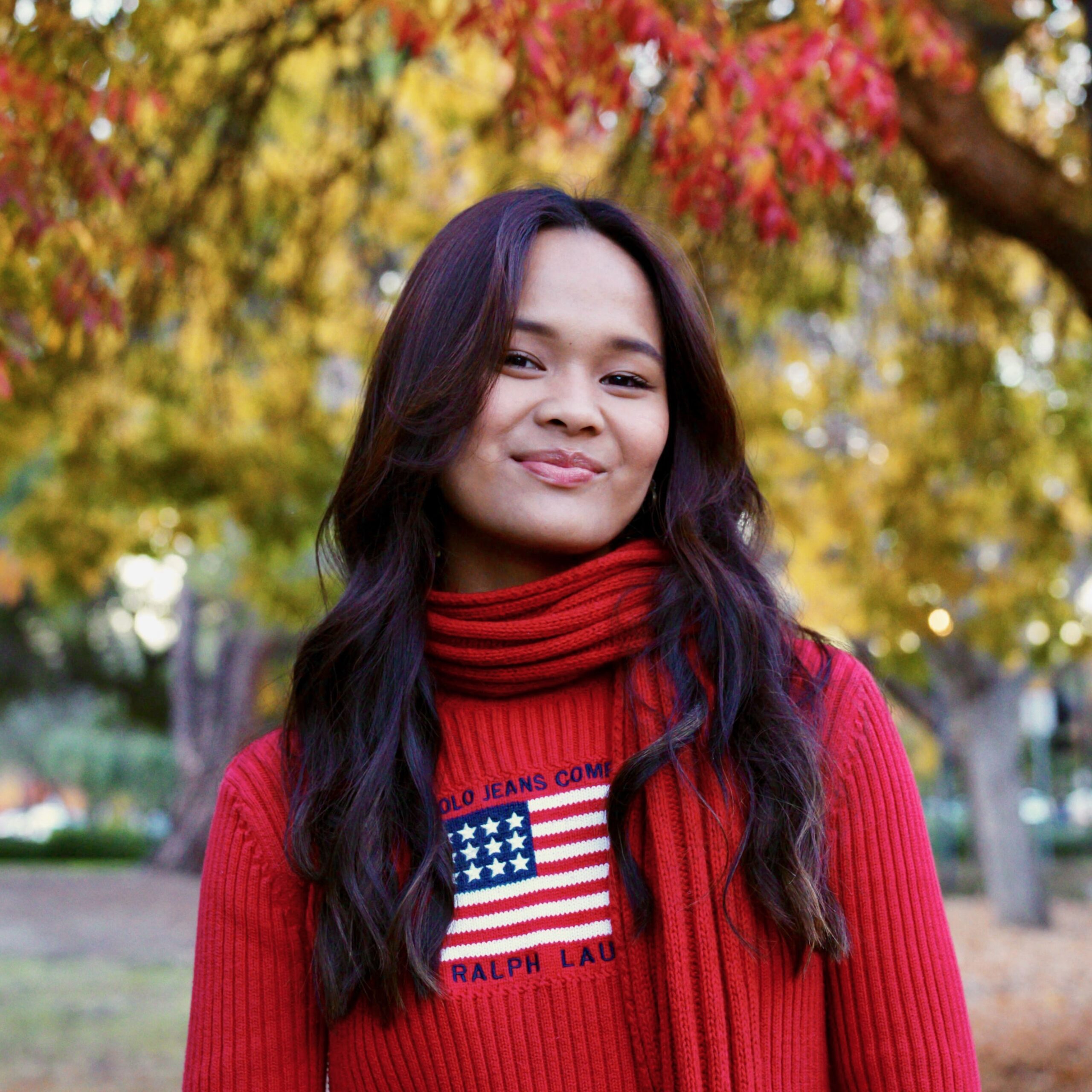 A photo of Isabella in a red sweater and red scarf in front of fall folliage