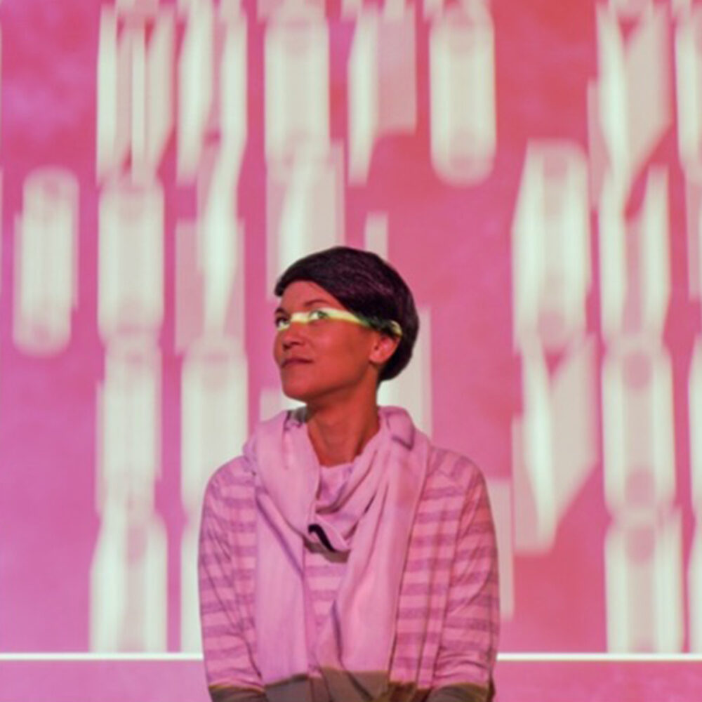 Lauren Lee McCarthy, a Chinese-American woman with short dark hair in front of blurred falling letters projected, a line of light through her eyes.