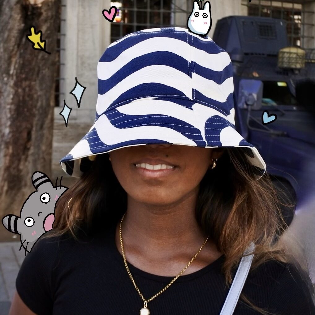 A photo of Nicole with a blue and white striped bucket hat pulled over her eyes. Doodles of tiny creatures peak out from behind her