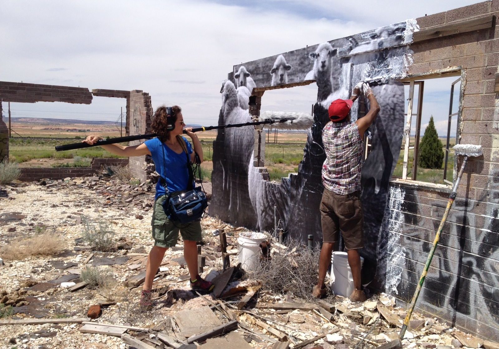 A documentary filmmaker is holding a boom mic to capture the audio of their film's subject who is painting a wall in the middle of a landscape.