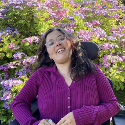 Photo of Sylvia in a purple blouse in front of a bush of bright purple flowers