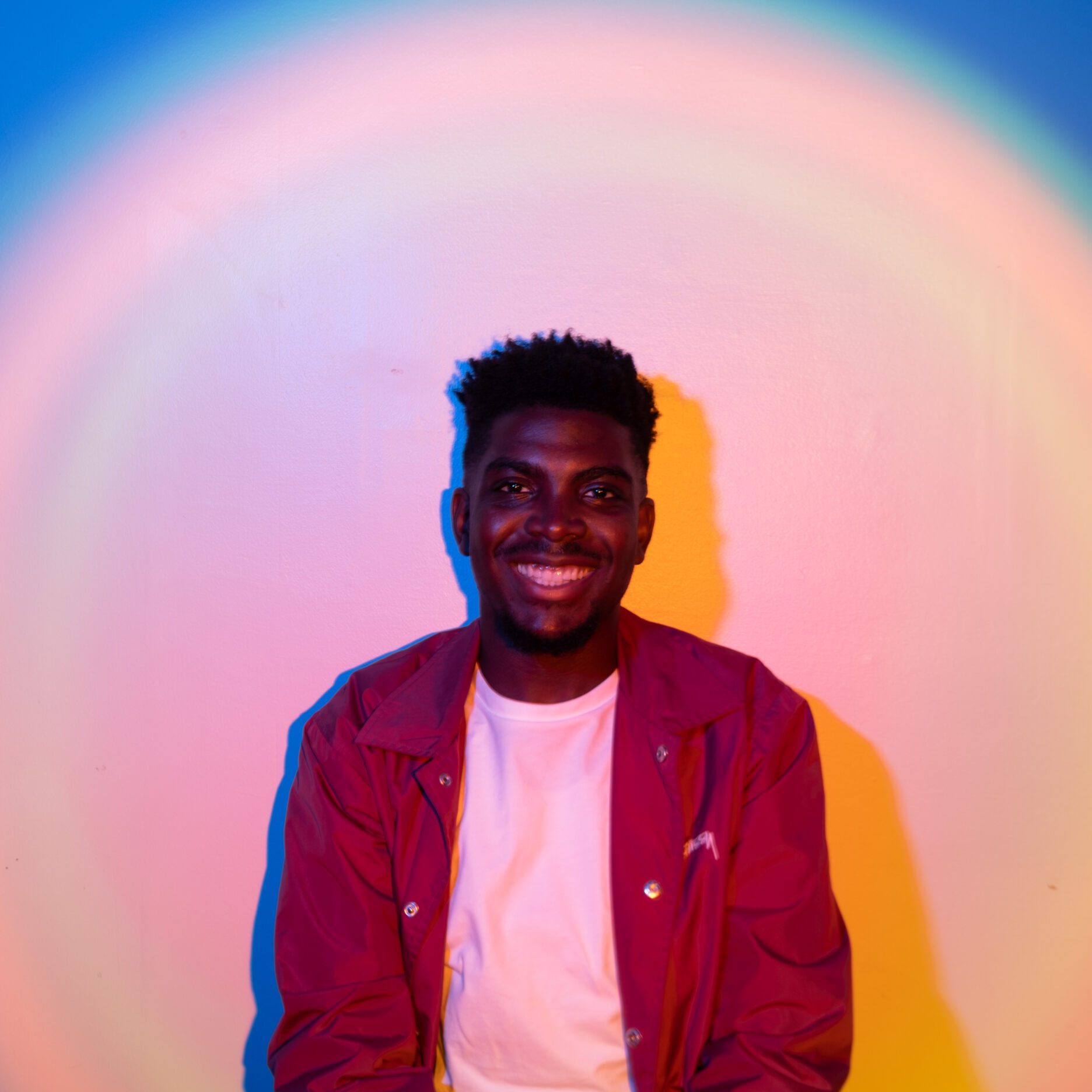 a photo of Tamilore Awosile wearing white shirt and red jacket. He's in a spotlight and the photo is edited to have bright rings circling the spotlight