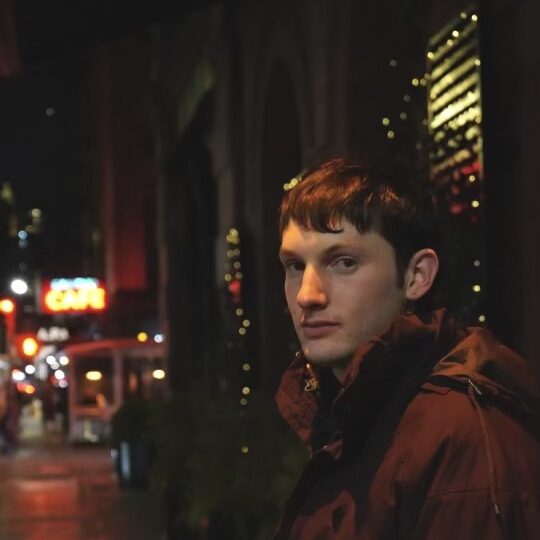a photo of Callum Tresnan wearing a winter coat on a dark street with blurred neon store fronts in the background