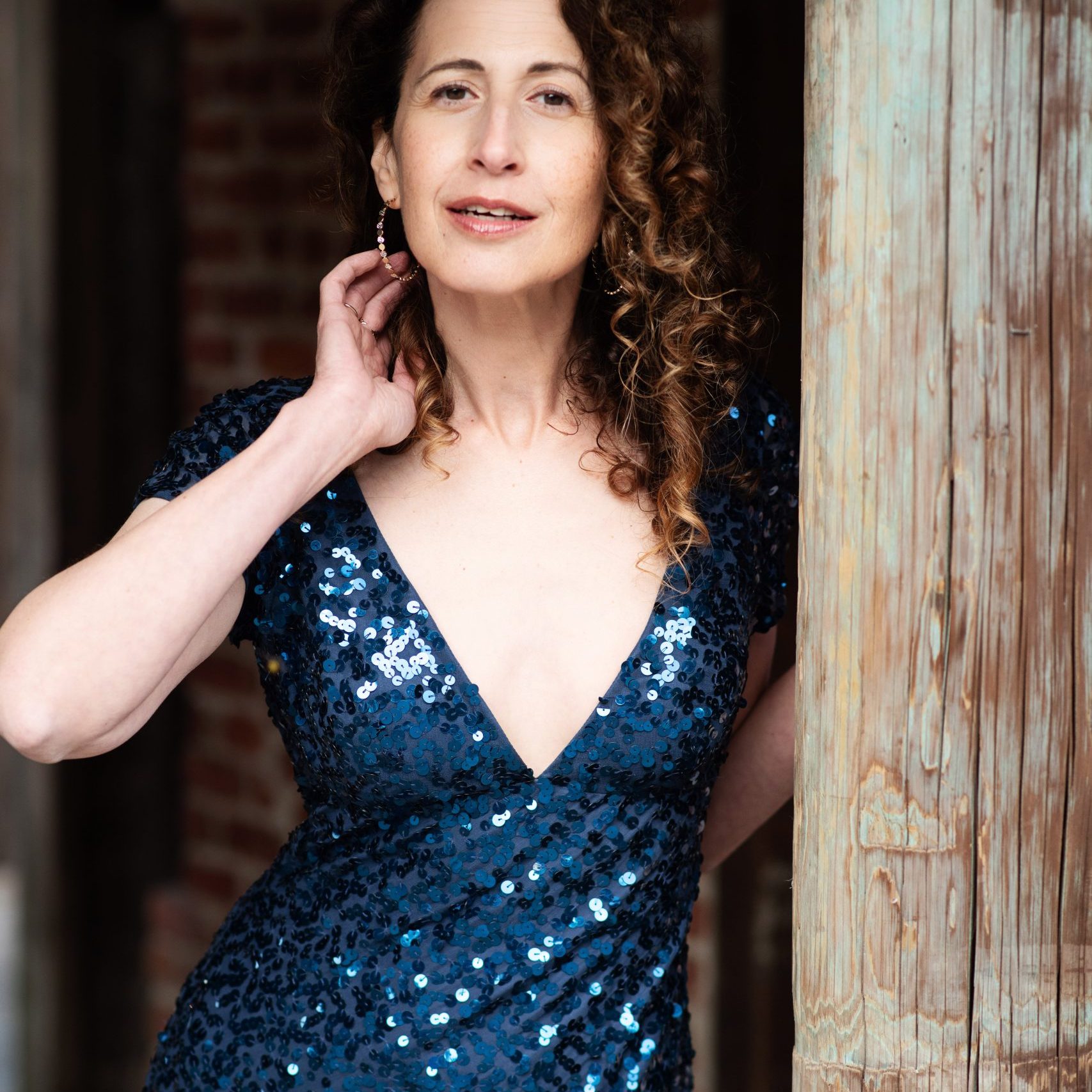 A person wearing a sparkly blue dress, standing in a doorway, smiling wistfully at the camera