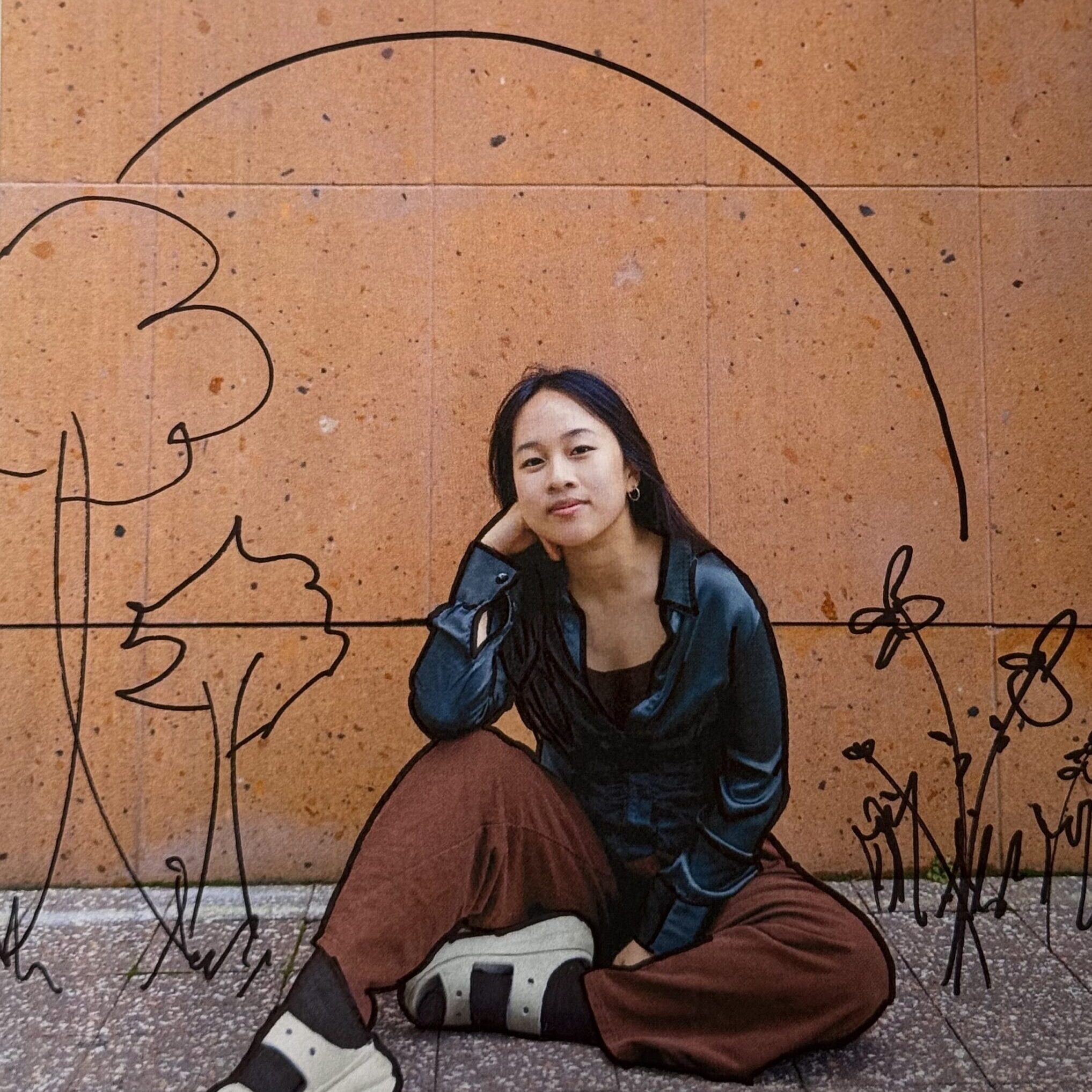 a photo of Junah sitting in front of an orange wall, with black doodles of trees and plants