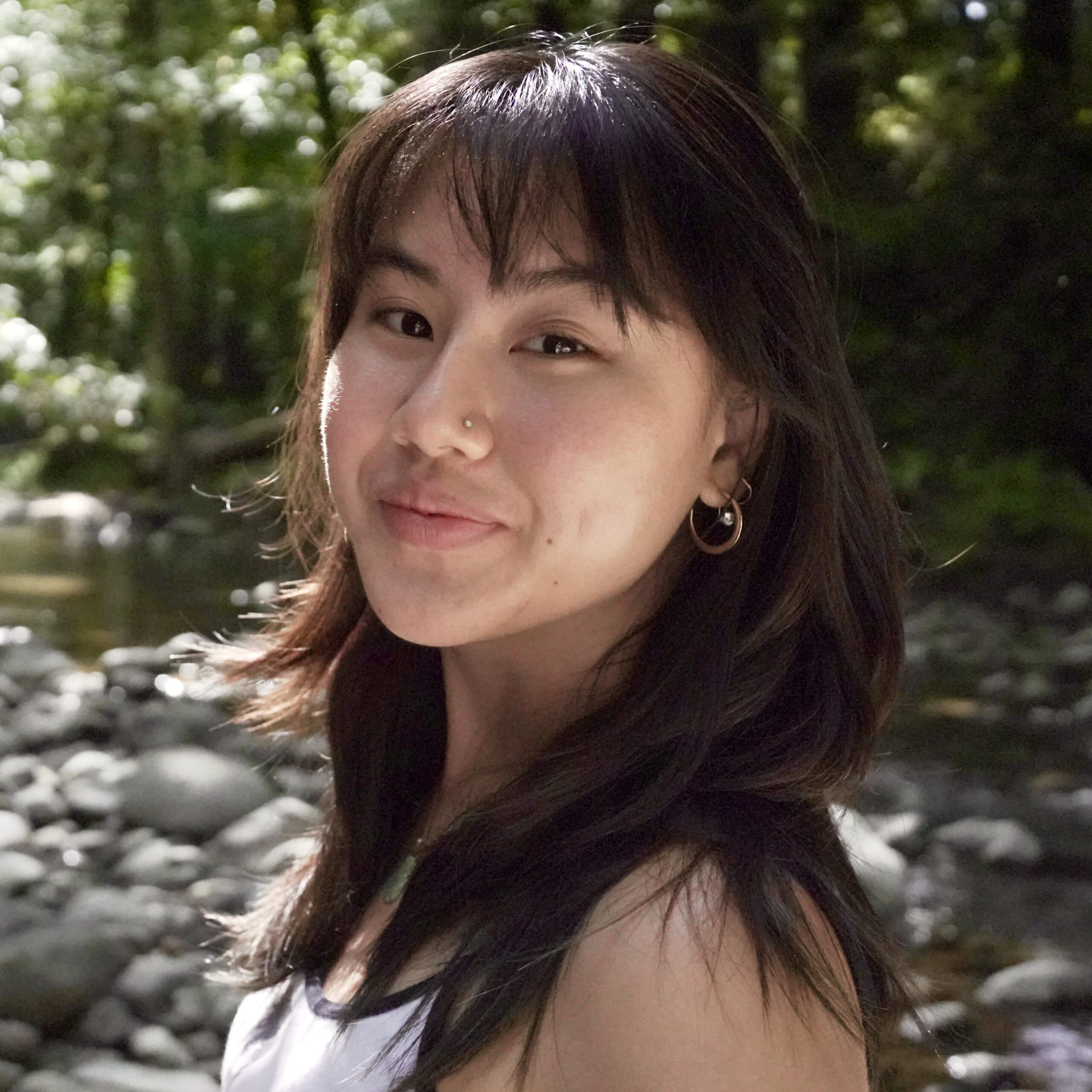 Portrait of Lora Suprandi: a young asian woman with long black hair in a forest with a stream